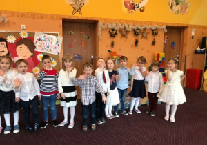 Grupa 8 odświętnie ubranych dzieci przesyła pocałunki oraz macha ręką do aparatu. Dzieci znajdują się w sali przedszkolnej. W tle widać tablicę z sylwetami uśmiechniętej babci oraz dziadka oraz plakat z napisem „Kochamy Was”.
