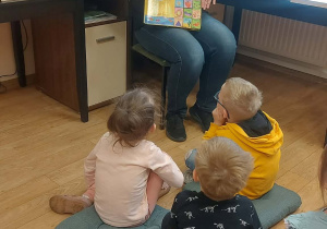 Dzieci siedzą na poduszkach i słuchają pani bibliotekarki