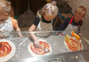 Przedszkolaki smarują ciasto na pizzę sosem pomidorowym.