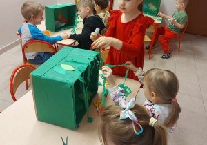 Dzieci podczas tworzenia przestrzennej pracy plastycznej z wykorzystaniem pudełka, bibuły przedstawiającej świat dinozaurów