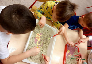 Dzieci za pomocą pędzli szukają na tackach wypełnionych piaskiem znalezisk prehistorycznych