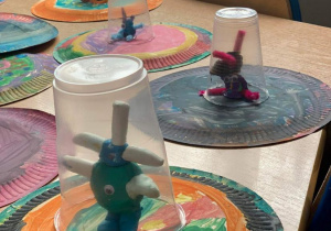 Prace plastyczne wykonane przez dzieci: kolorowe statki kosmiczne wykonane z pomalowanego farbą talerzyka papierowego, ufoludka z plasteliny oraz plastikowego kubeczka