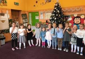 Grupa 8 odświętnie ubranych dzieci recytuje wiersze. Dzieci znajdują się w sali przedszkolnej. W tle widać ubraną choinkę bożonarodzeniową oraz tablicę z sylwetami uśmiechniętej babci oraz dziadka.