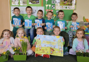 Zdjęcie grupowe dzieci trzymające napis wiosna z wiosennymi kwiatami w sali przedszkolnej