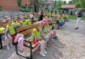 Dzieci siedzące w ogrodzie biblioteki i słuchające opowiadania czytanego przez pracownika biblioteki