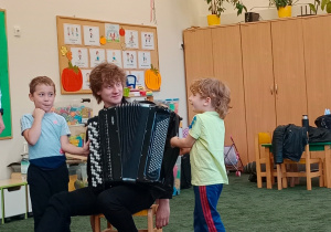 Muzyk z akordeonem siedzi na krześle a dzieci próbują grać na jego instrumencie muzycznym