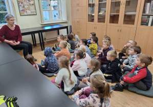 Dzieci siedzą na poduszkach w bibliotece i słuchają pracownika