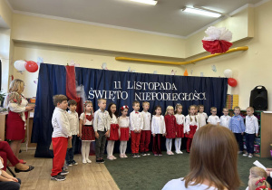 Grupa dzieci ubranych w stroje barwy biało-czerwone na tle dekoracji o charakterze patriotycznym