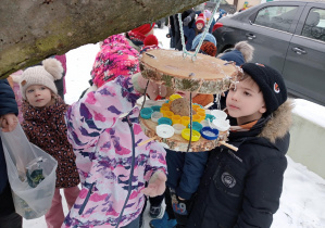 Zdjęcie przedstawia dzieci oglądające karmnik dla ptaków zawieszony na drzewie.