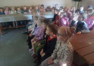 Dzieci siedzą przy stolikach i słuchają.