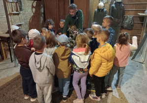Dzieci stoją dookoła stolika, na którym znajdują się różne przedmioty i słuchają przewodnika