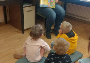 Dzieci siedzą na poduszkach i słuchają pani bibliotekarki