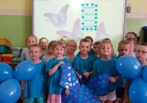 Wspólne zdjęcie dzieci z grupy 2. Dzieci ubrane są na niebiesko, trzymają wyciętego motyla. W tle widać napis ,,Jesteśmy z Wami Niebieskimi motylami"