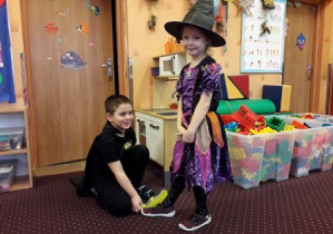 Zdjęcie przedstawia dwoje dzieci podczas jednej z zabaw karnawałowych. Chłopiec kuca obok dziewczynki. Trzyma papierową sylwetę pantofelka a dziewczynka stoi i uśmiecha.